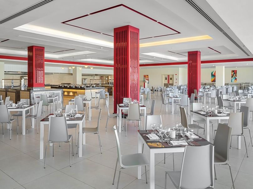 Empty dining area with white tables, chairs, and red columns, and a counter in the background.