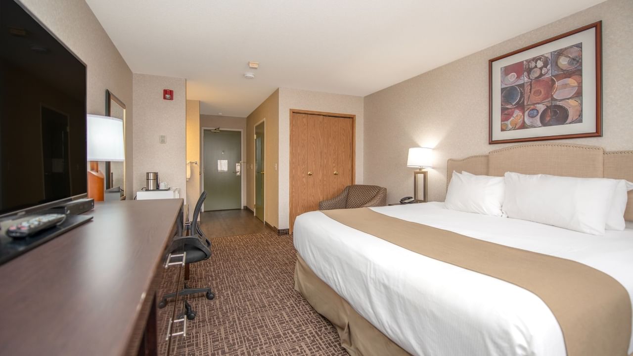 A hotel room with a bed, desk, chair, TV, and artwork on the wall.