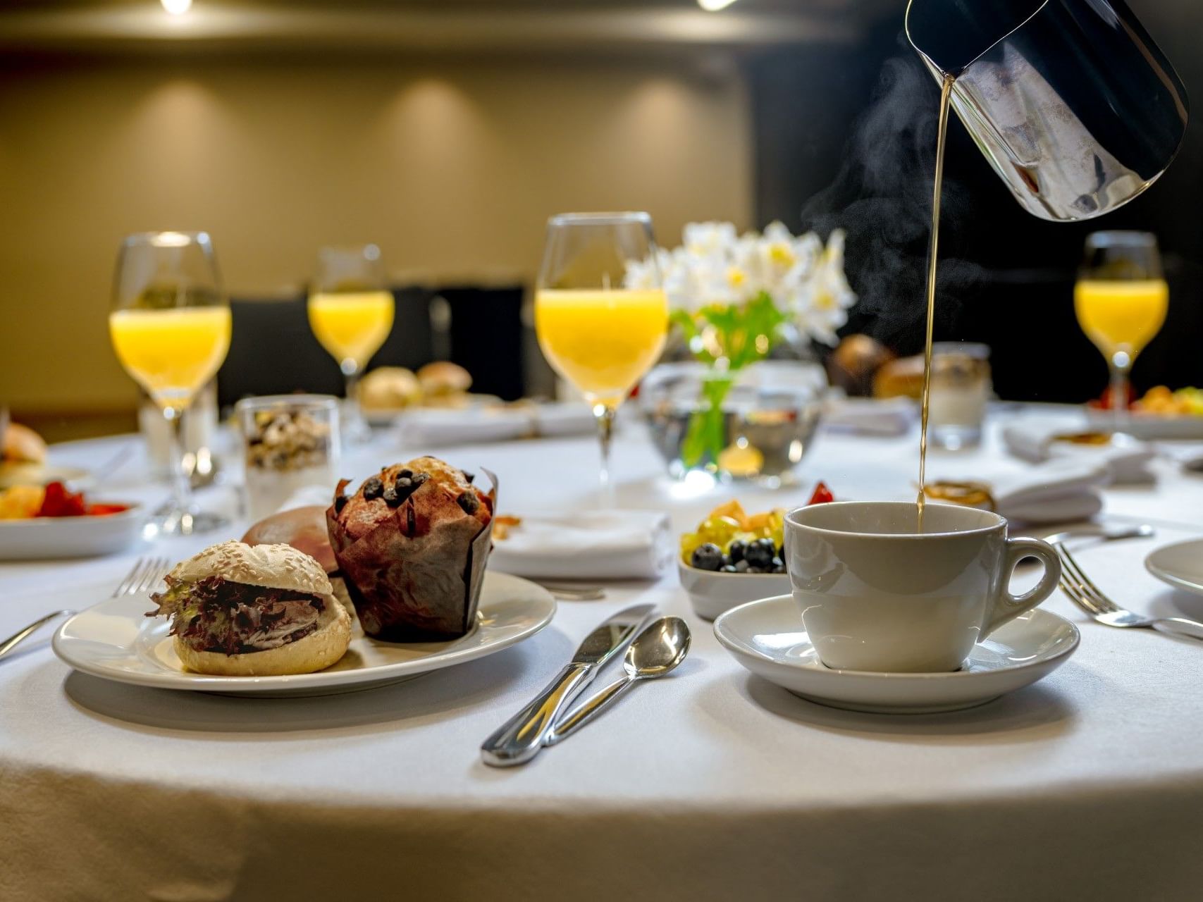 Se está sirviendo café en una taza en una mesa de desayuno en la que hay diversos alimentos y cubiertos.