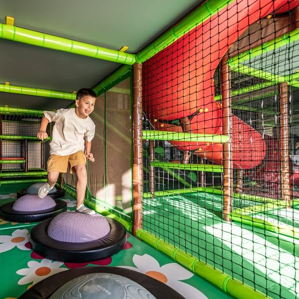 Young boy standing on exercise ball in colorful indoor play area with large red slide and green net.