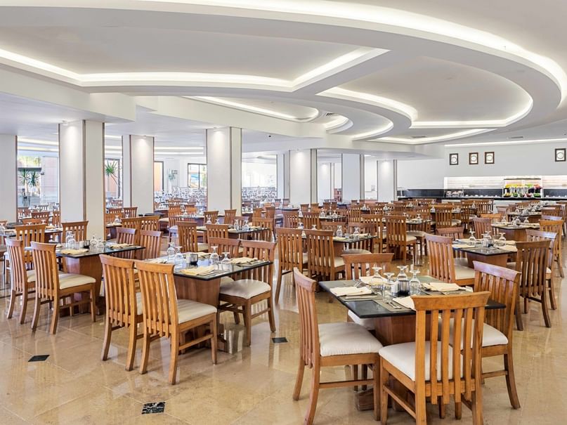 Large, empty dining hall with rows of wooden tables and chairs, curved ceiling lights, and a counter in the background.