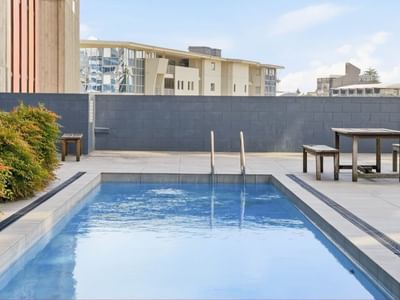 Outdoor pool with wooden table and benches surrounded by greenery at Student Living Auckland - Beach.