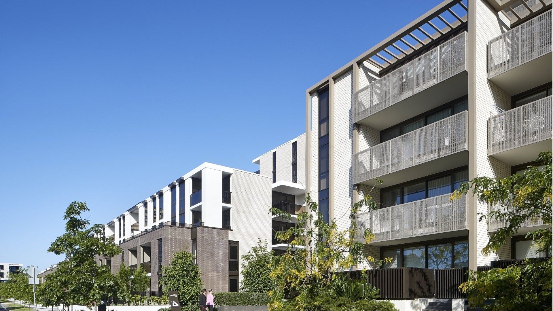 Modern apartment complex with balconies, surrounded by trees and greenery under a clear blue sky.