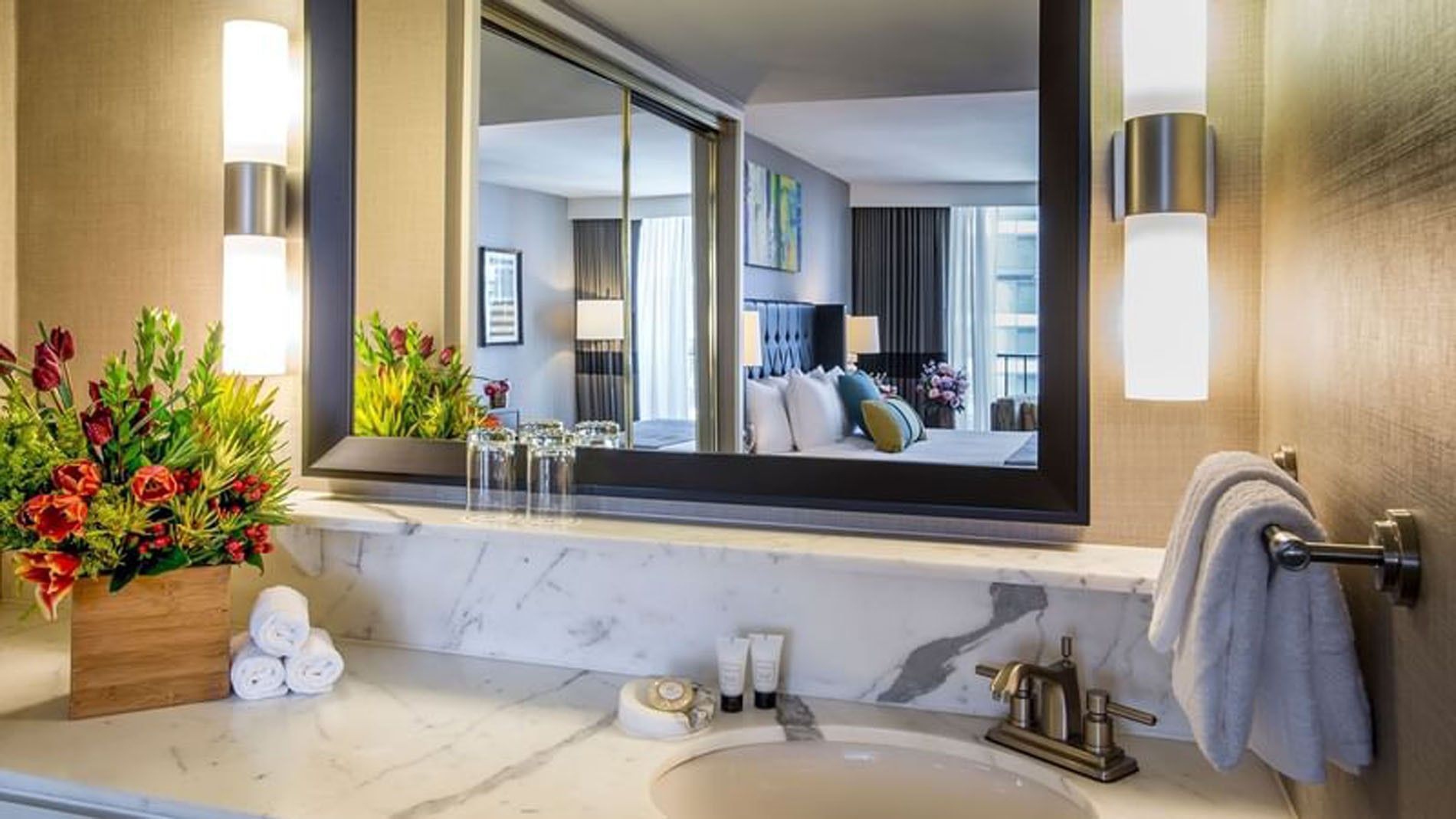 A hotel bathroom vanity with a large mirror, marble countertop, flowers, and towels.