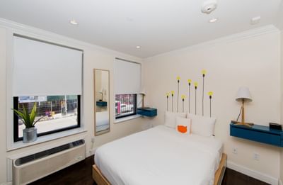 East Village Hotel bedroom with white walls, bed, blue shelving, yellow light accents, potted plant, and air conditioner.