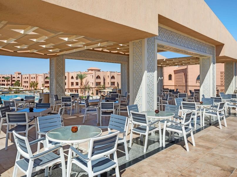 Outdoor dining area with tables and chairs under a canopy, overlooking a pool and resort buildings.