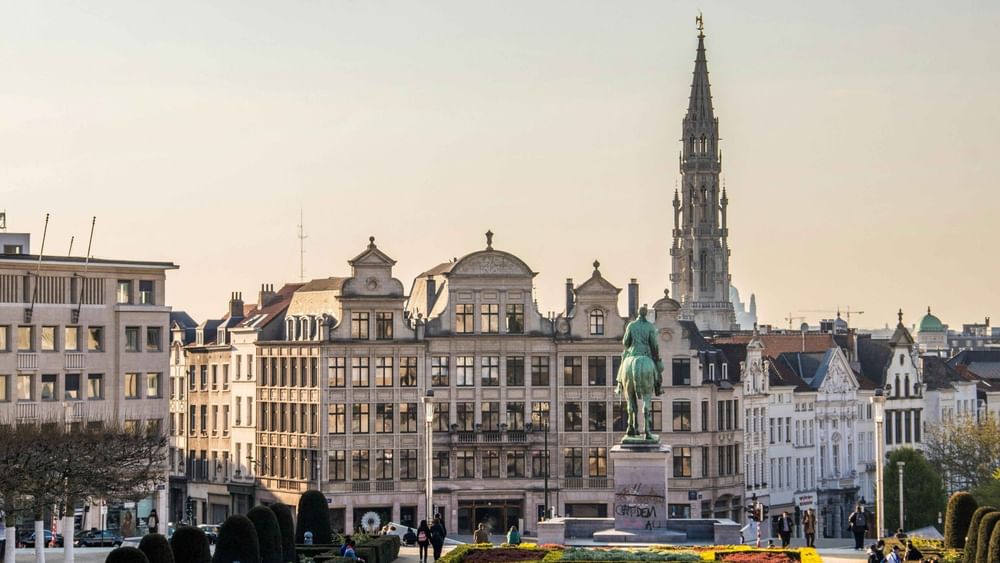 Historic Brussels cityscape with ornate buildings, a bronze statue, colorful gardens, and a Gothic spire under soft evening light.