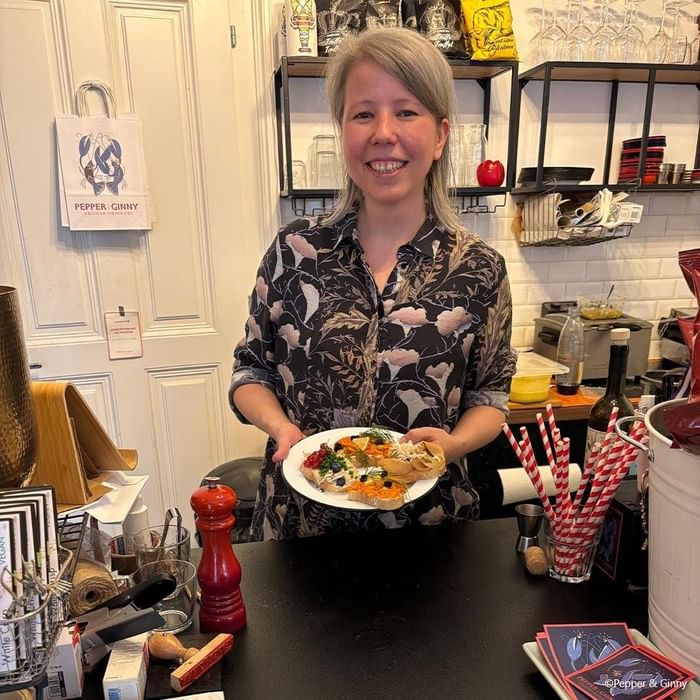 Frau mit hellgrauem Haar hält Teller mit Speisen in vegetarischem Restaurant, Tür im Hintergrund.