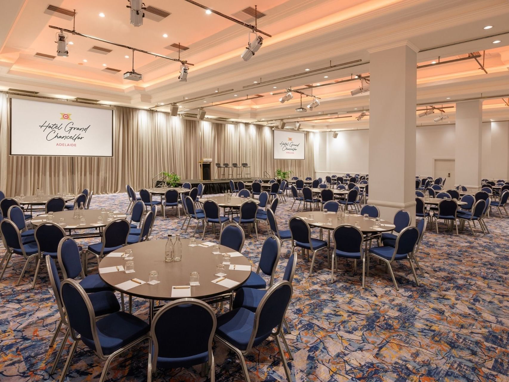 Banquet hall setup with round tables and chairs under a large screen advertising The Complete Conference Experience.