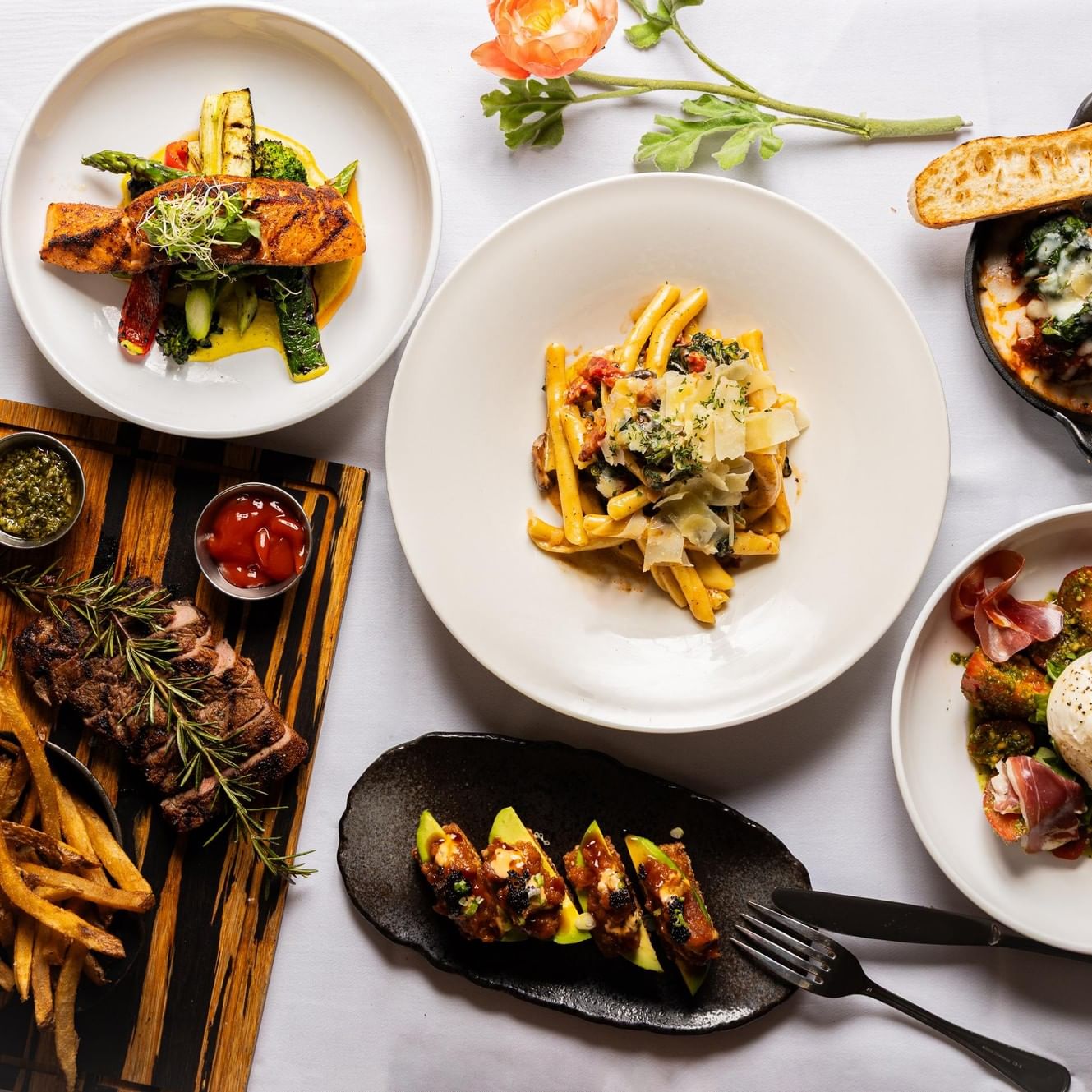 Overhead view of diverse gourmet dishes including grilled salmon, pasta, sliced steak with fries, and burrata with prosciutto