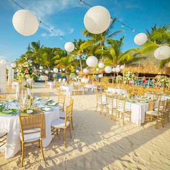 Beach venue with golden chairs, white tables, and hanging lanterns, surrounded by palm trees at Paradise Beach Hotel