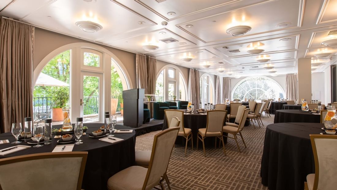 La Terraza Ballroom at El Prado Hotel, set with round black tables, chairs, and a stage with arched windows