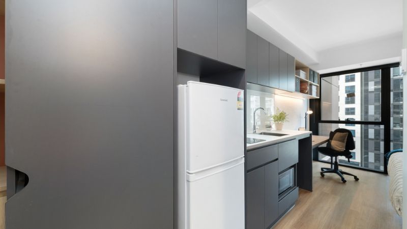 Modern kitchenette with gray cabinets, white fridge, sink, and desk with chair by large window.