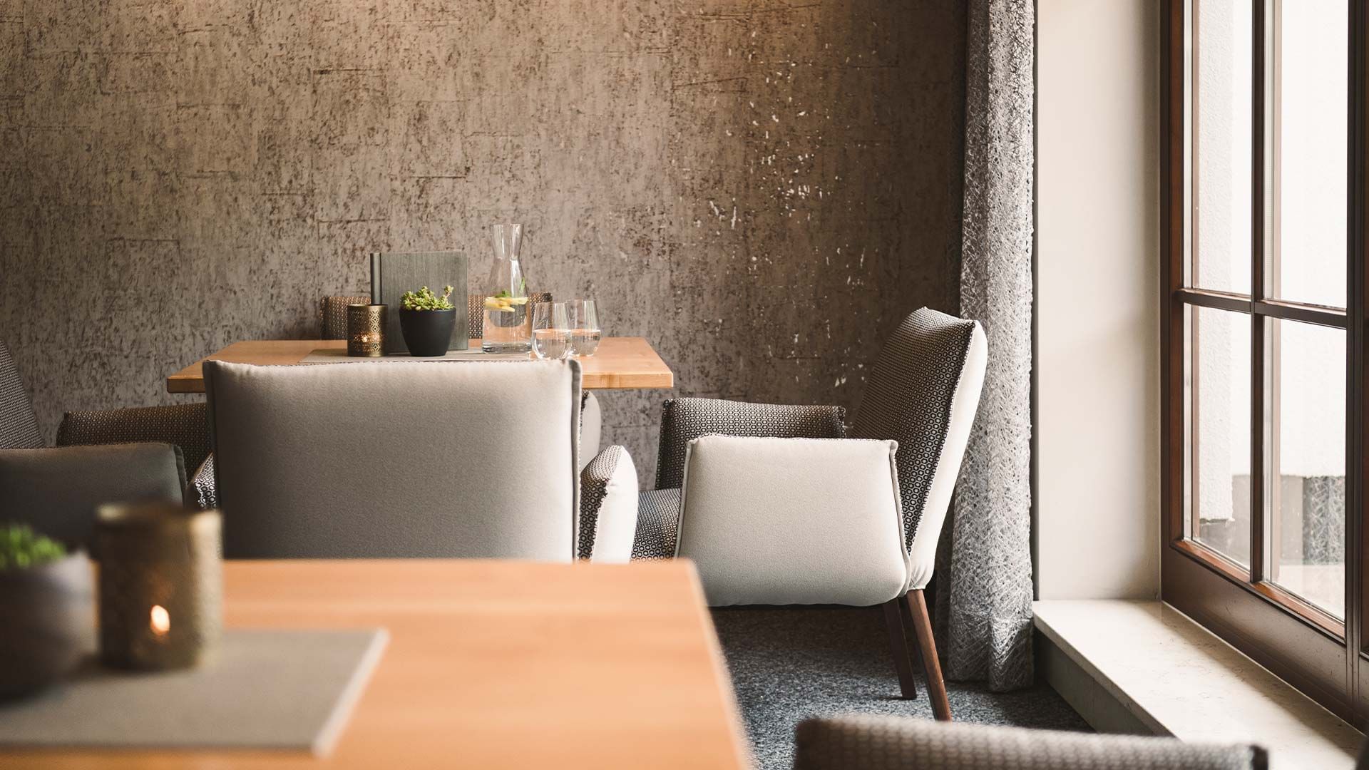 A dining area with wooden tables, cushioned chairs, a glass pitcher, and a window with curtains.