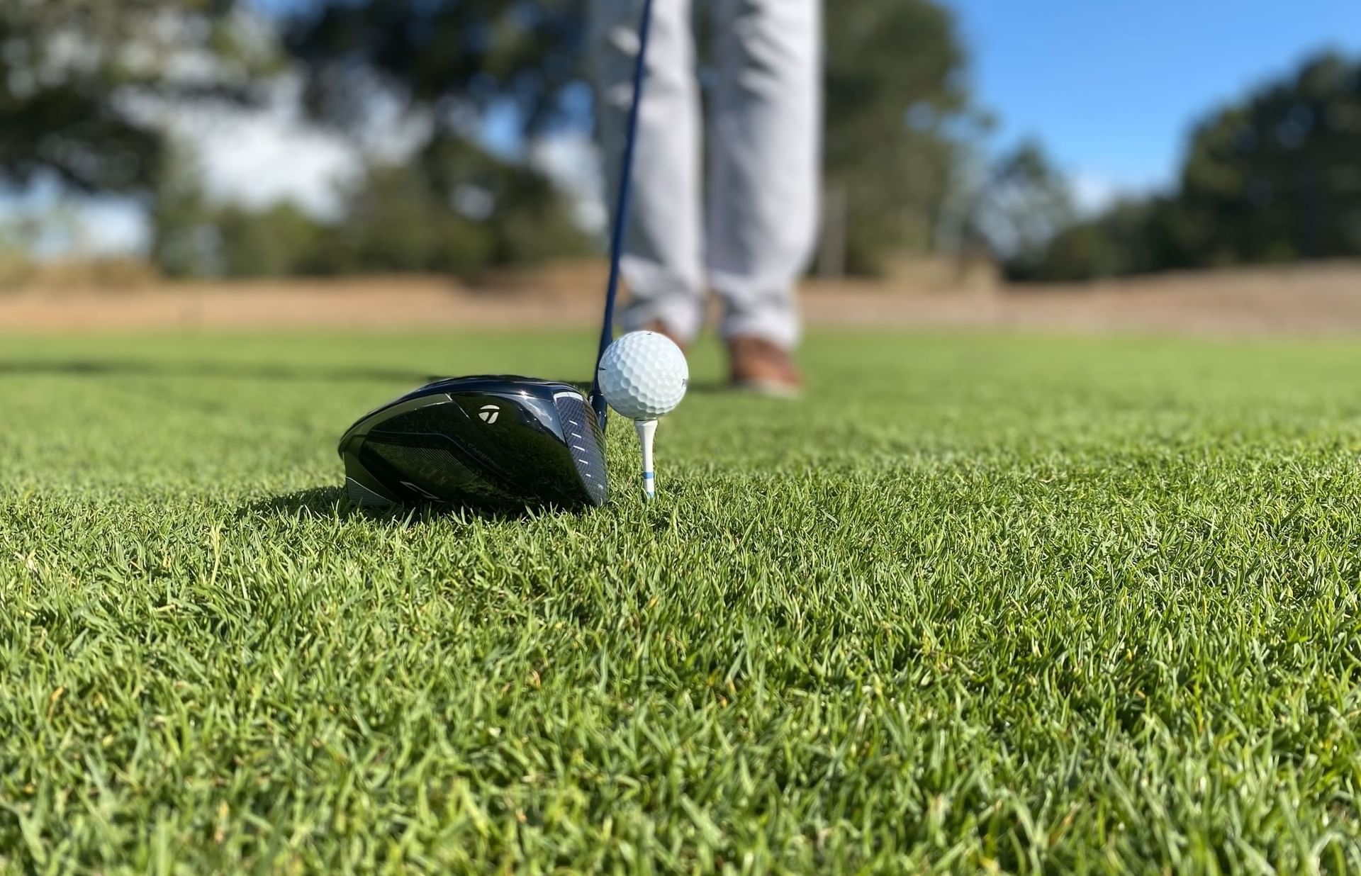Golfer with club positioned to hit a golf ball on green grass, with trees in the background.