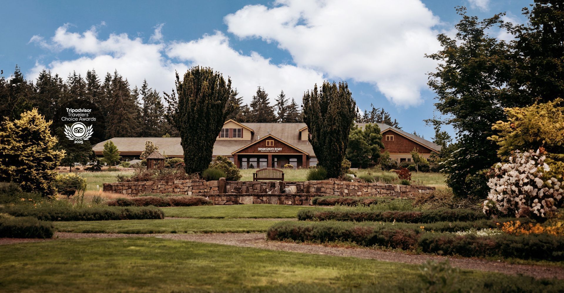 A scenic house with a lawn, stone wall, and a bench in front, surrounded by trees and flowers.