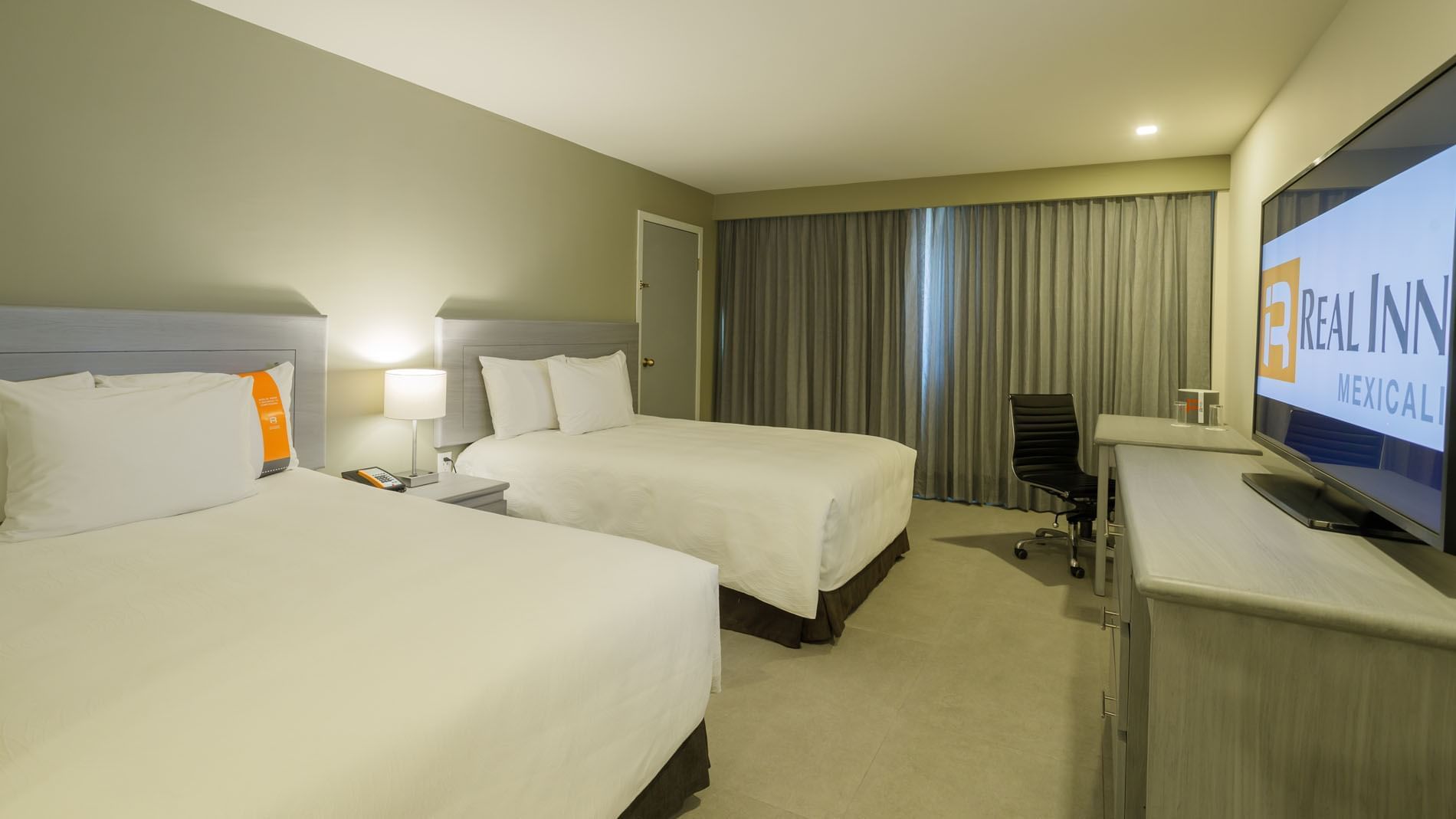 2 Double Deluxe room with white linens, a sleek black desk chair, and grey blackout curtains at Real Inn Mexicali