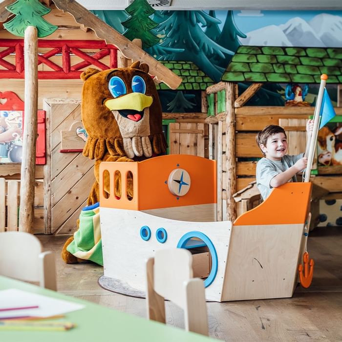 Ein Junge spielt in einem Spielzimmer neben einem Maskottchen in einem Holzboot.