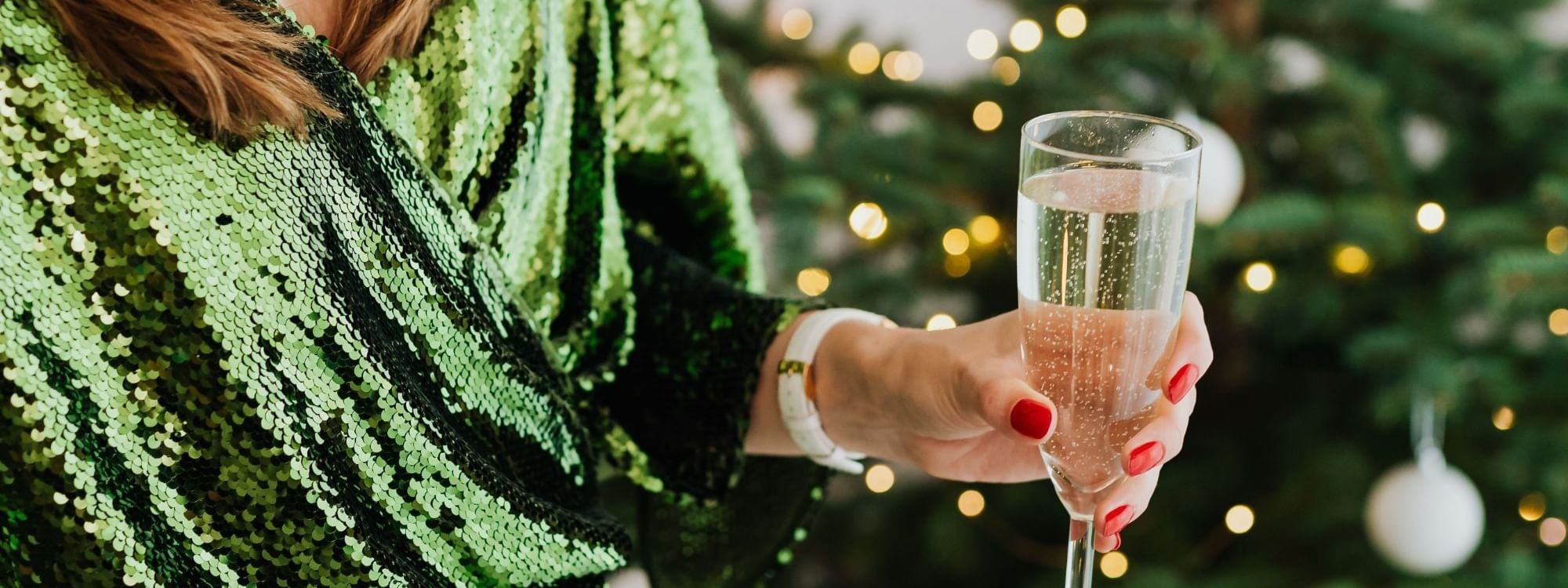 Woman in green sequined top holding champagne glass with decorated Christmas tree and twinkling lights in background
