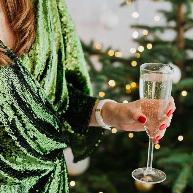 Woman in green sequined top holding champagne glass with decorated Christmas tree and twinkling lights in background