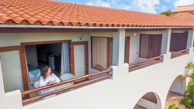 Smiling woman with a mug standing on a white balcony under a tile roof outside the Garden View Room at Sugar Cane Club