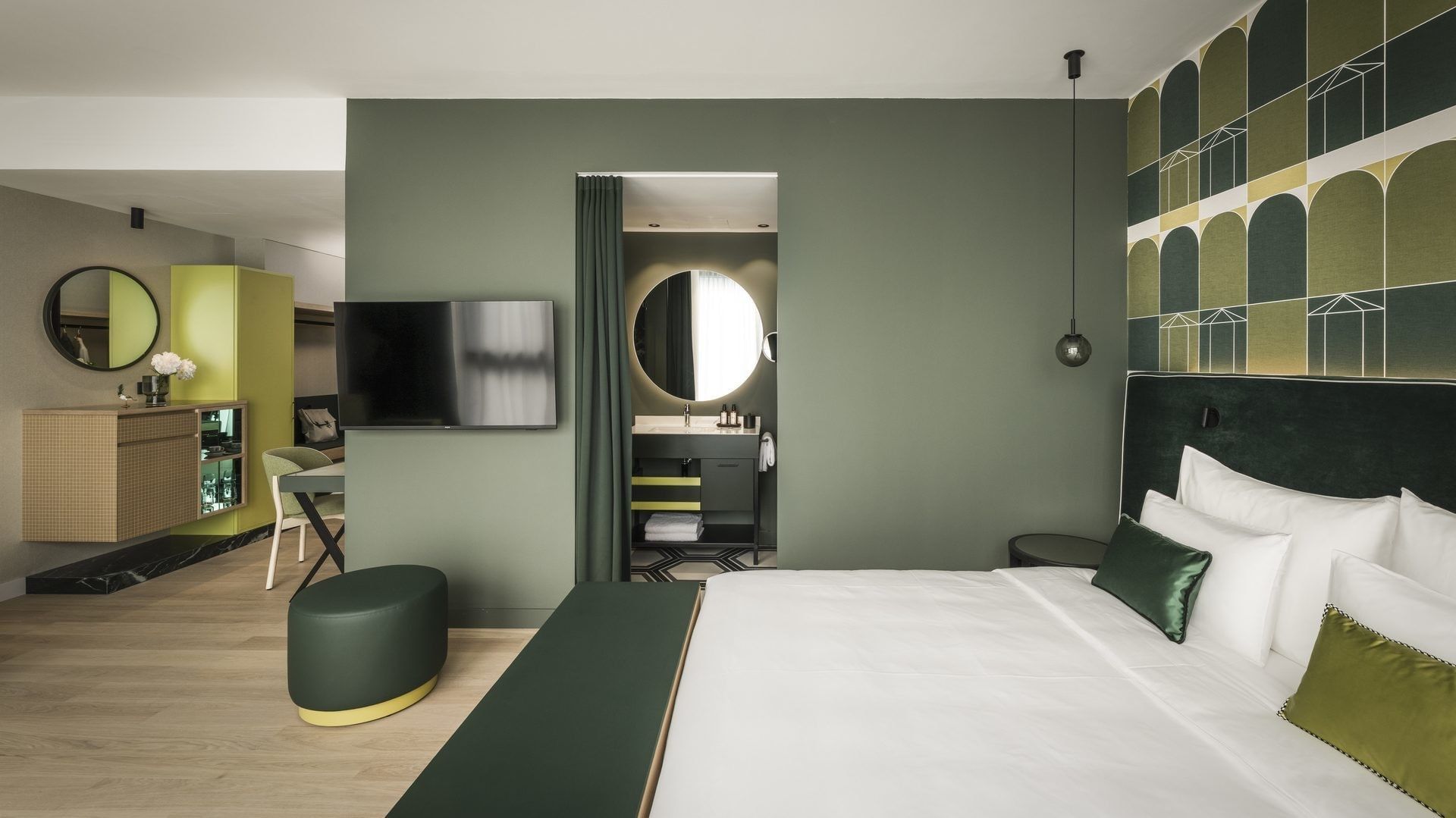 Grünes Hotelzimmer mit Bett, Fernseher, Spiegel, Kommode, Hocker und sichtbarem Badezimmer.