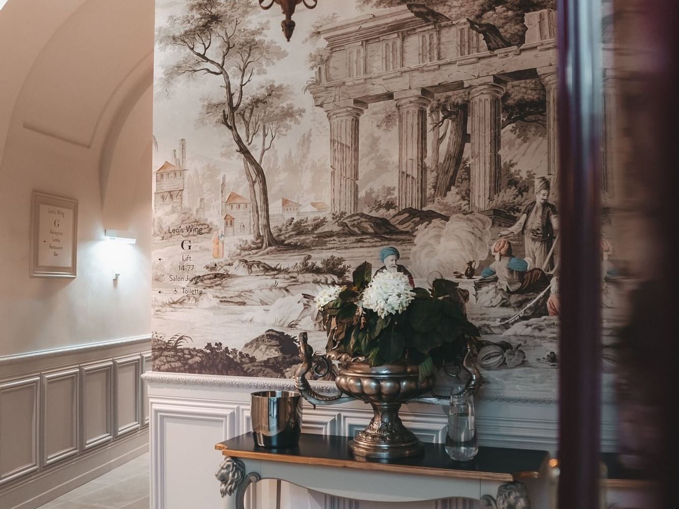 Hallway with ornate chandelier, mural, and table with vase and flowers near door.