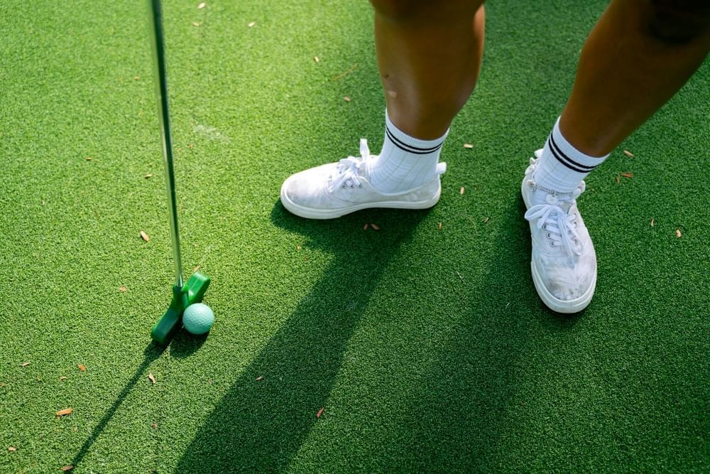 A close-up of feet wearing white sneakers and high socks standing on turf beside a mini golf club and a green golf ball.
