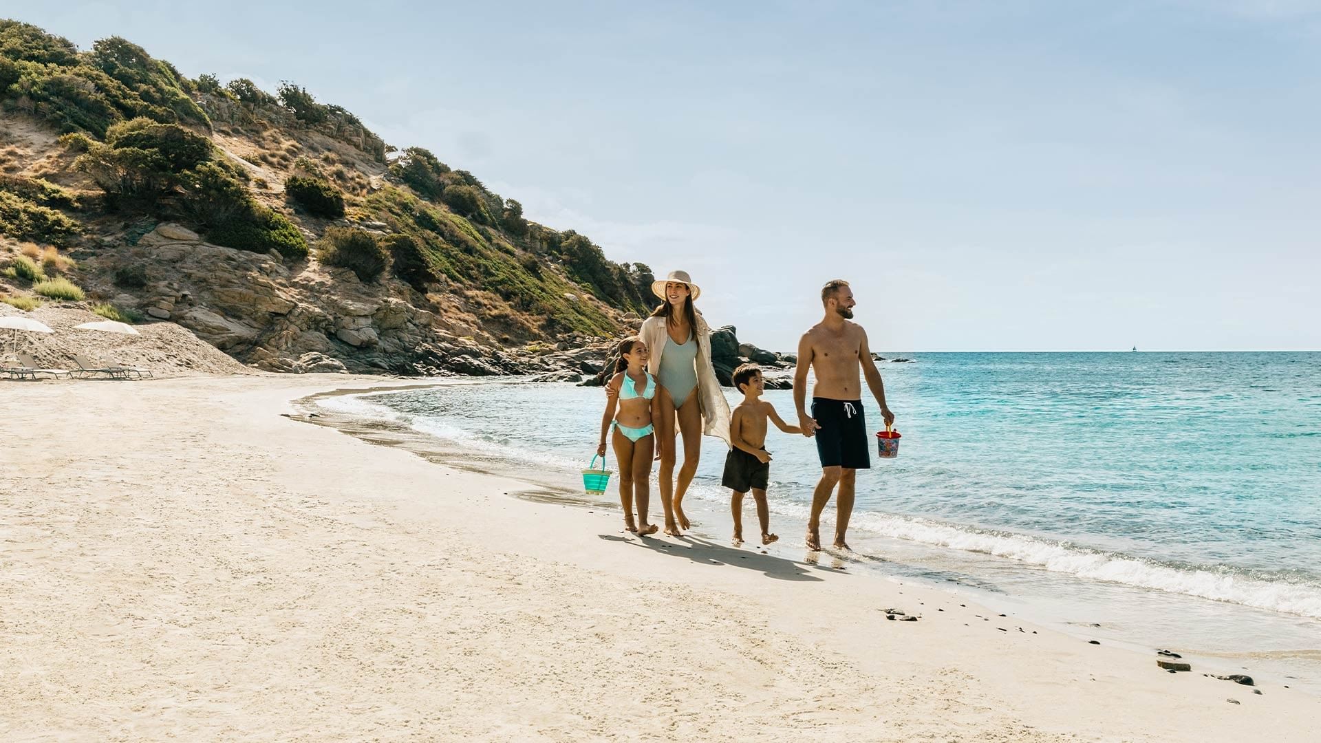 Family of four walks together on beach with blue ocean and rocky hill in background.