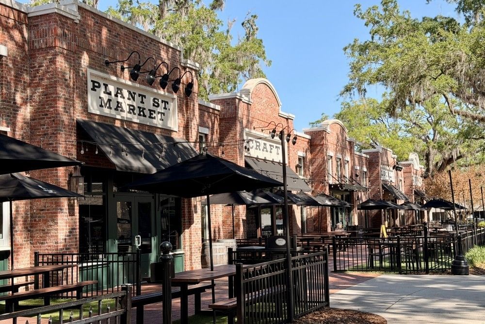 A brick building with a sign that reads Plant St. Market with black awnings, umbrellas, and many picnic tables on an outdoor patio.