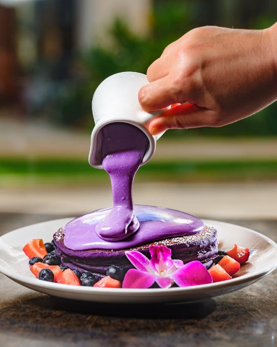 a hand pouring purple syrup on a stack of purple pancakes with fruit and a flower
