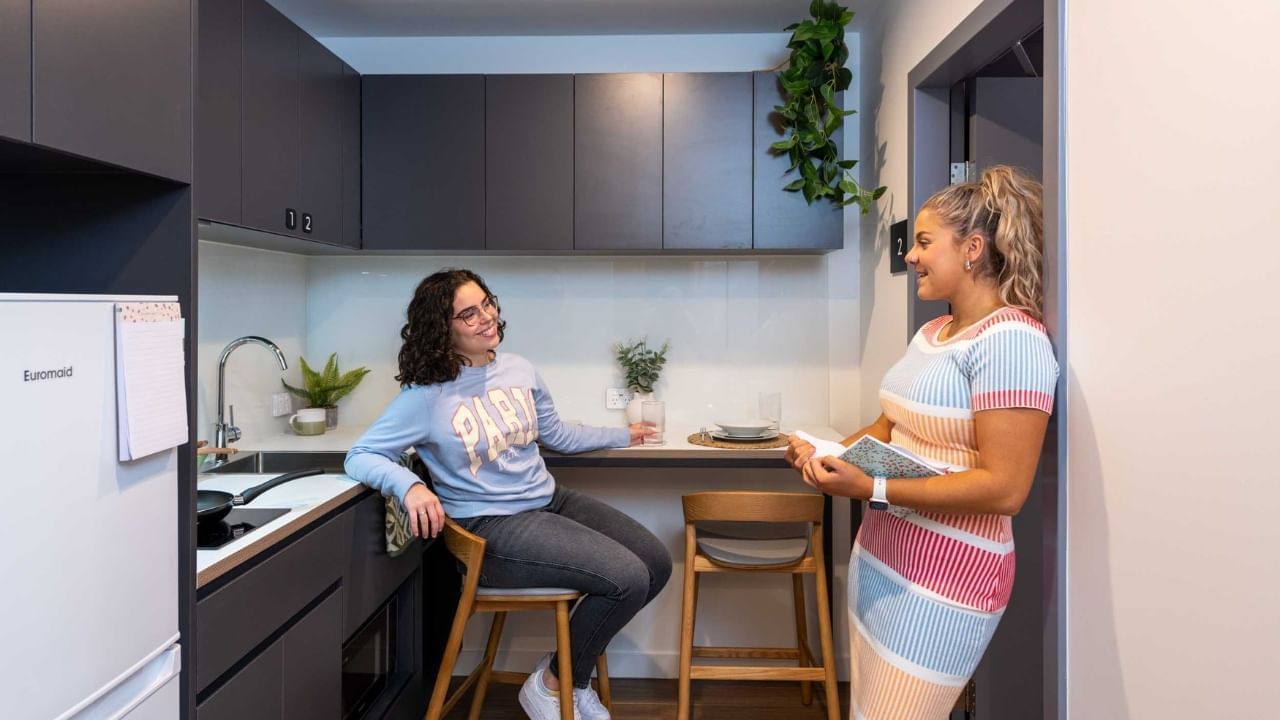 Two residents in a small kitchen, one seated on a stool, the other holding a book.