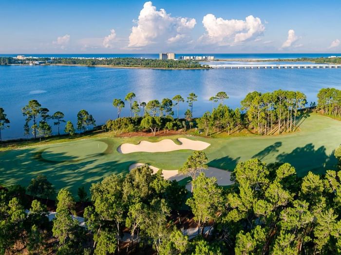 Aerial view of Shark's Tooth golf course hole #15 with sand bunkers surrounded by pine trees along a coastal bay with blue waters.