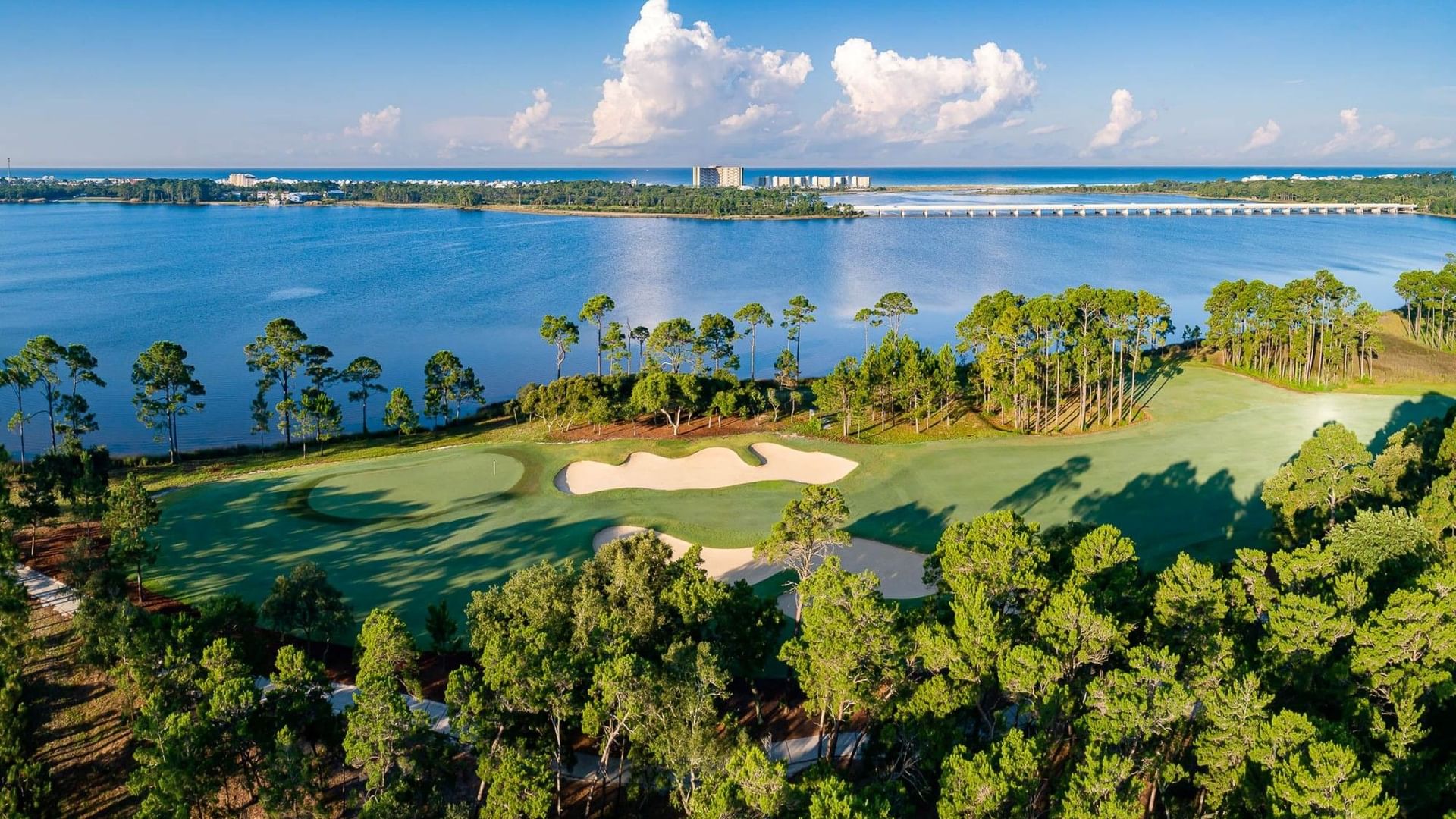 Aerial view of Shark's Tooth golf course hole #15 with sand bunkers surrounded by pine trees along a coastal bay with blue waters.