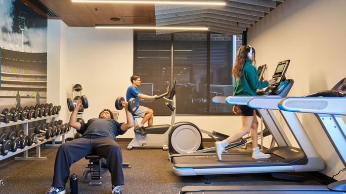People exercising with dumbbells, a weight bench, and treadmills at UniLodge Royal Melbourne in North Melbourne.