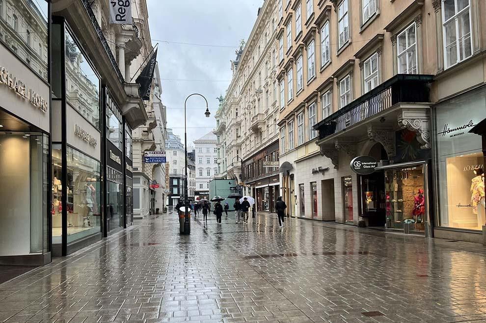 Einkaufsstraße in Wien bei Regen mit Boutiquen, Passanten und spiegelndem Pflaster.
