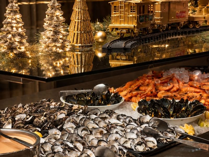 Festive seafood buffet with oysters, prawns, and mussels displayed beside illuminated decorative trees and a golden train.