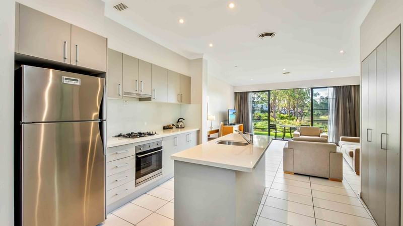 Kitchen with stainless steel appliances open to a living room with a view of greenery outside at Mercure Kooindah Waters