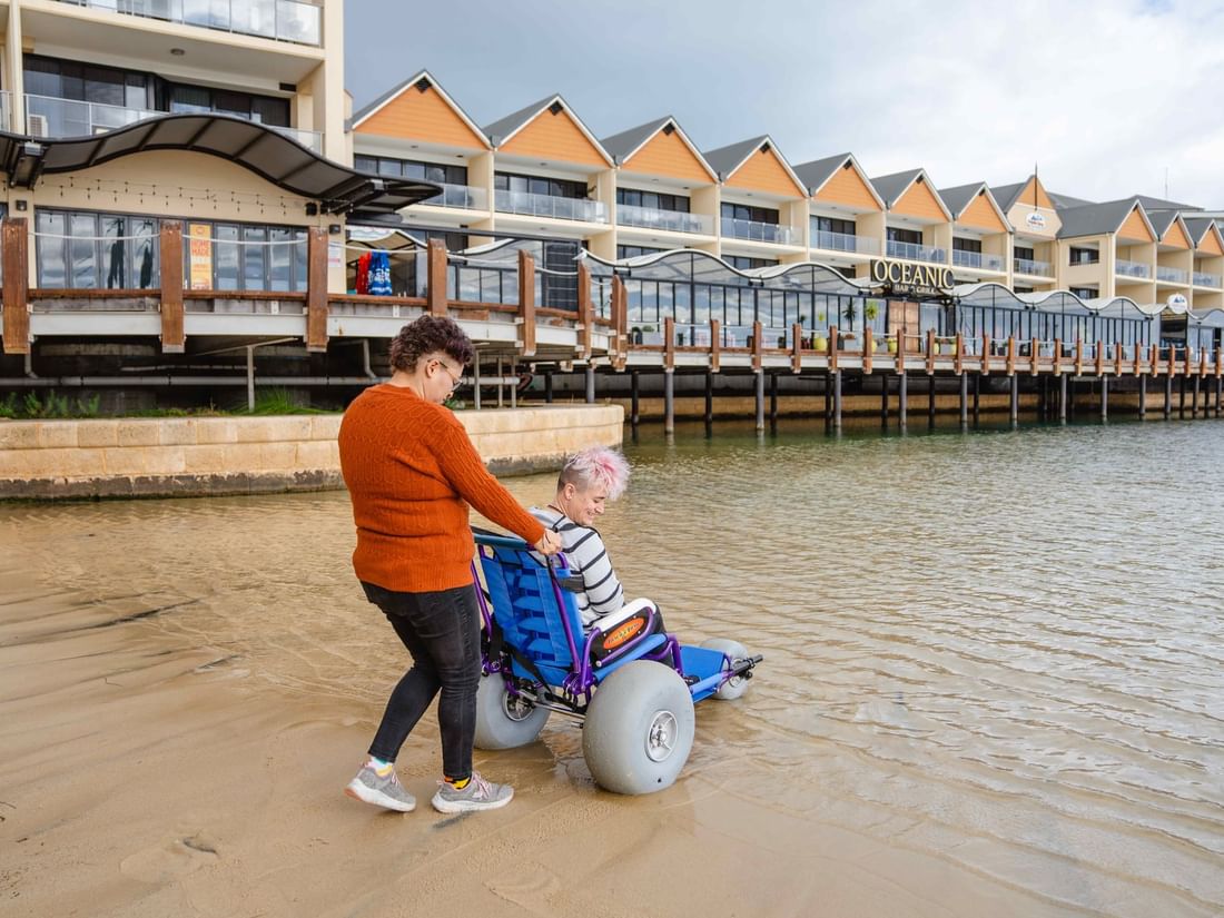 Woman pushes a person in a large-wheeled beach wheelchair on the sand near The Sebel Mandurah