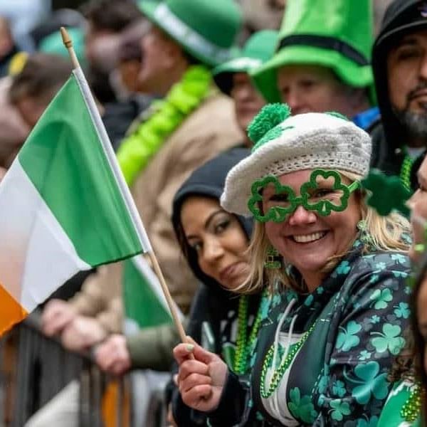 Smiling woman in St Patrick's Day parade with Irish flag and crowd behind her.