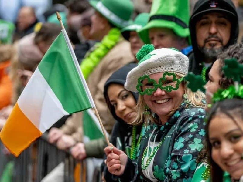Smiling woman in St Patrick's Day parade with Irish flag and crowd behind her.