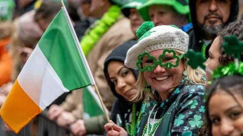 Smiling woman in St Patrick's Day parade with Irish flag and crowd behind her.