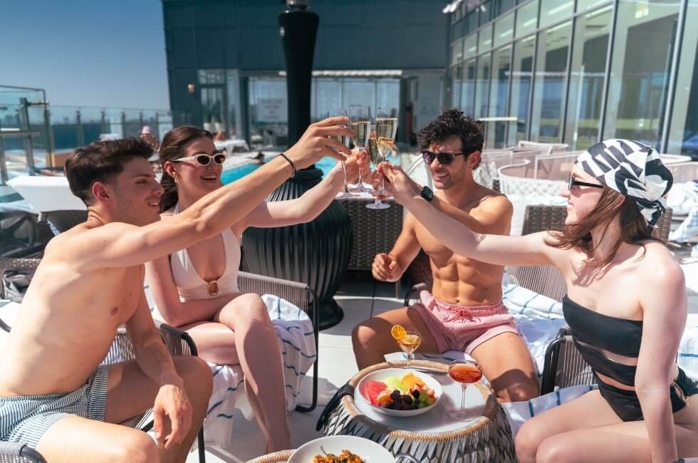 Four guests toast drinks by poolside lounge chairs under sunny skies on the outdoor rooftop terrace at Hotel X Toronto