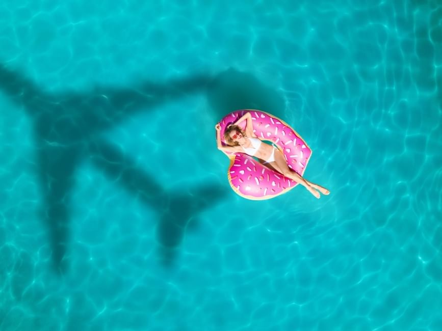 Aerial view of a woman relaxing on a pink donut float in the clear pool at Fiesta Inn with a plane’s shadow overhead