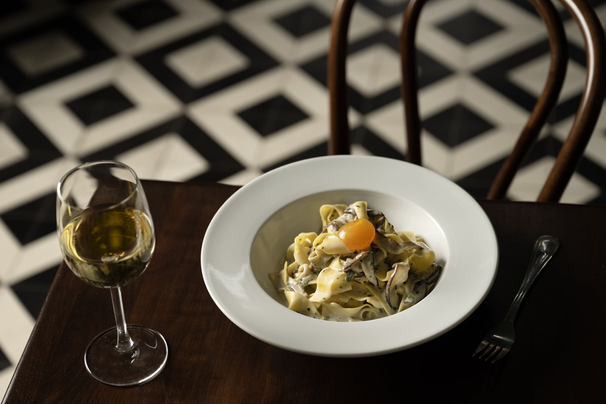 A plate of Coco Miso Pappardelle with shiitake mushroom, lemongrass, cured egg yolk on a table with a wine glass