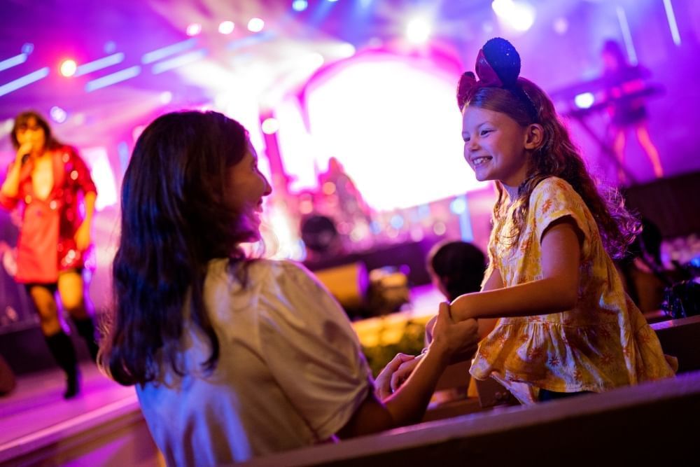 A woman holding a girl's hand in front of a purple lit stage with a performer out of focus onstage in the background.