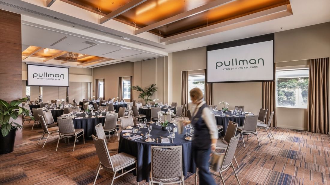 Spacious event room with round tables, elegant place settings, and Pullman branding on projection screens.