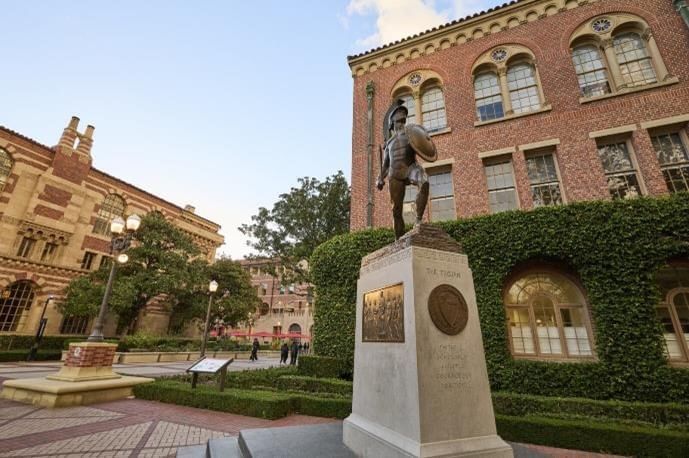 A statue stands in front of a building with a hedge and a paved area.