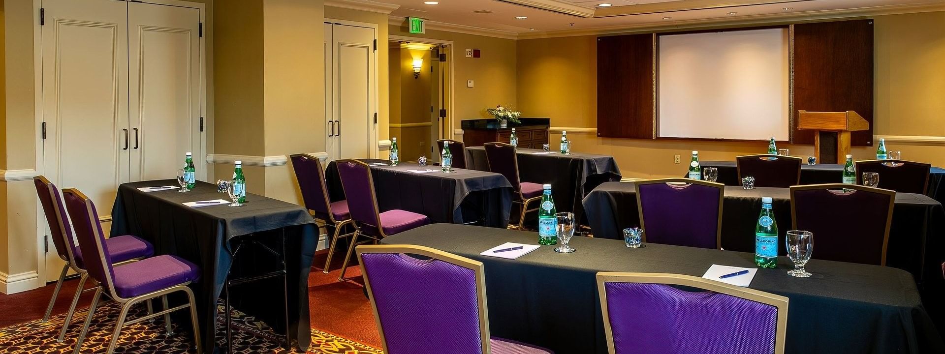 Purple chairs by black tables under a projector screen near a wooden lectern in Tabor II at Warwick Denver