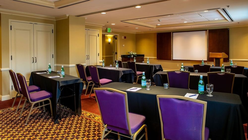 Purple chairs by black tables under a projector screen near a wooden lectern in Tabor II at Warwick Denver